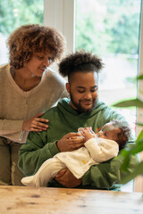 Poster - Mother looking at father feeding baby daughter at home