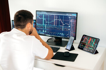 Concentrated male trader sitting at desk at home office monitoring stock market looking at monitors analyzing price flow, risks and prospects. Stock trading, cryptocurrency concept