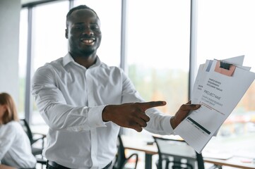 Wall Mural - Black man is standing and holding documents. Group of office workers are together indoors