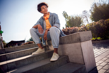 Canvas Print - Photo of positive cheerful dreamy youngster with afro hairdo dressed jeans jacket in headphones sitting on stairs in park outdoors