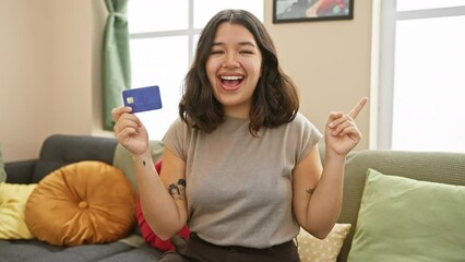 Canvas Print - Portrait of a beaming young hispanic woman on her sofa at home, cheerfully pointing to the side with her credit card in hand, exuding confidence and joy