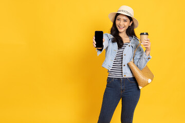 Happy Young Asian traveler woman showing blank screen mobile phone and holding hot coffee cup isolated on yellow background, Tourist concept