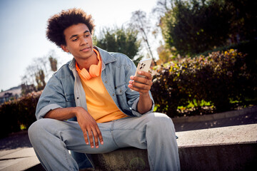 Canvas Print - Photo of good mood youngster with afro hairdo dressed jeans jacket headphones sitting on platform texting email on smartphone outdoors