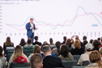 Wall Mural - Blurred speaker discussing chart trends to audience at corporate seminar. Focus on engaged attendees.