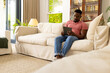 © Wavebreak Media - African American man in a pink shirt and jeans is focused on a tablet while sitting on a cream sofa