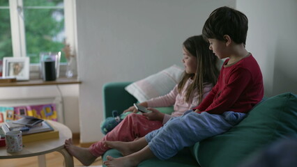 Wall Mural - Little brother and sister looking at cellphone screen while seated on couch sofa in living room. Children hypnotized by content online sharing gadget