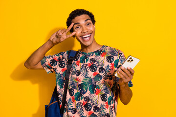 Wall Mural - Photo of friendly guy student with rucksack show v sign cover face symbol victory holding smartphone isolated on yellow color background