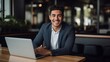 © Wajid - Capture the essence of a good-looking millennial office employee or student sitting at a desk in front of a laptop, smiling, and confidently looking at the camera.