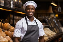 Happy Black Baker Man In A Black Apron And A White Chef's Hat On His Head On The Background Of A Bread Counter