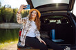 © maxbelchenko - Selfie in nature. Happy woman sits in the trunk of a car and uses a smartphone. The concept of traveling by car, active lifestyle, blogging.