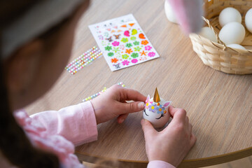 A cute girl with pink bunny ears makes an Easter craft - decorates an egg in the form of a unicorn with rhinestones, horn, flowers in the interior of a house with plants.