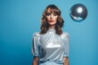 © katrin888 - A stylish young woman in retro outfit and makeup poses confidently near a mirror ball on a blue background. She wears a silver sparkly top with balloon sleeves and high-waisted pants, exuding glamour.