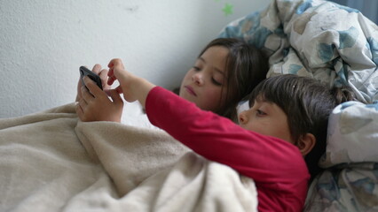 Wall Mural - Kids lying in bed looking at cellphone device, sister sharing phone screen with smaller brother watching entertainment media content online, children absorbed by technology