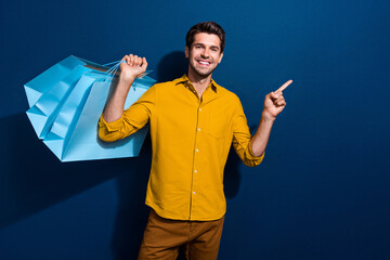 Poster - Photo portrait of attractive young man shopper bags point empty space dressed stylish yellow clothes isolated on dark blue color background