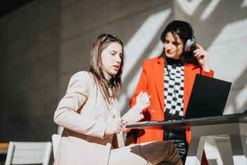 Wall Mural - Two focused businesswomen engaging in a collaborative work session outdoors, using a laptop in a well-lit, urban setting.