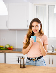 Wall Mural - Young woman in casual clothes standing near kitchen table with cup of tea or coffee at home