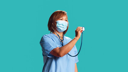 Wall Mural - Asian experienced nurse doing mock up showing of properly using professional stethoscope. Knowledgeable clinic worker demonstrating medical gear usage, isolated over blue studio background