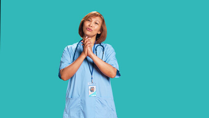 Wall Mural - Pleading asian healthcare professional imploring hospital management for wage raise. Overworked nurse with hands folded in prayer gesture asking for days off, isolated over blue studio background