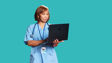 Wall Mural - Focused asian nurse typing patient data on laptop after finishing medical checkup appointment. Clinic employee writing informations on gadget, isolated over blue studio background