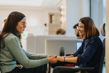 Cheerful young women employees engaged in a moment of solidarity and teamwork, holding hands across a table in an office setting, embodying support and partnership.