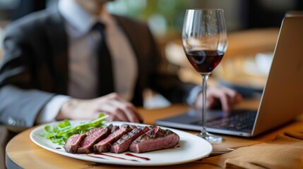 A person is sitting at a table working on a laptop. On the table next to the laptop is a plate with a steak cut into slices on it  