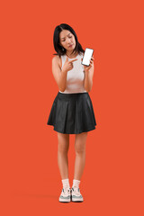 Poster - Thoughtful young beautiful Asian woman pointing at mobile phone on orange background