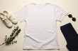 A plain white t-shirt is laid out flat next to casual white sneakers, dark sunglasses, and navy blue