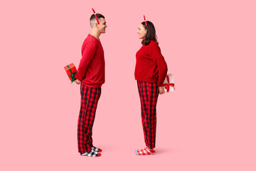 Wall Mural - Happy young couple in Christmas pajamas hiding gifts behind back against pink background