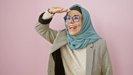 Wall Mural - Joyful middle age hispanic woman in hijab happily looking far away, hand shielding eyes. delighted in seeking beyond, against isolated pink background.