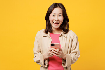 Wall Mural - Young surprised shocked woman of Asian ethnicity wear pink t-shirt beige shirt pastel casual clothes hold in hand use mobile cell phone isolated on plain yellow background studio. Lifestyle portrait.