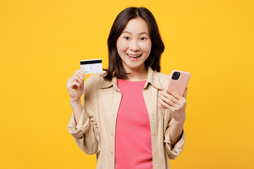 Wall Mural - Young woman of Asian ethnicity wear pink t-shirt beige shirt casual clothes using mobile cell phone hold credit bank card shopping online order delivery book tour isolated on plain yellow background.