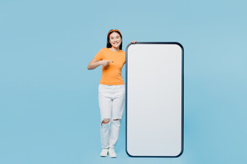Wall Mural - Young woman of Asian ethnicity wear orange t-shirt casual clothes point finger on big huge blank screen mobile cell phone smartphone with workspace copy space mockup area isolated on blue background.