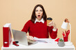 Young employee business woman wear red sweater sit work at office desk with pc laptop hold paper account documents drink coffee isolated on plain pastel beige background. Achievement career concept.