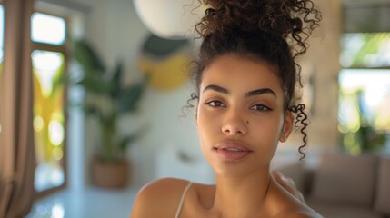 Wall Mural - A woman with curly hair and a side-swept bang wearing a white top looking directly at the camera with a soft expression set against a blurred indoor background with natural light.