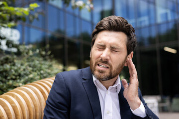 Close-up photo of a young businessman writhing in pain, sitting on a bench near the office and holding his hand by his ear