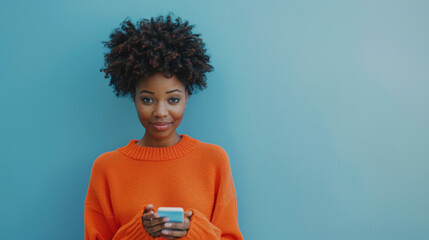 Wall Mural - A person in a bright orange sweater is engaged with a smartphone, standing against a solid blue background.