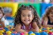 © DachAI - A joyful little girl with curly hair lies in a ball pit, smiling at the camera, with friends in the background