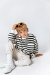 Little smiling blonde kid boy in striped sweatshirt ,white trousers and sunglasses sitting and posing at studio