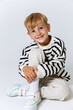 Little smiling blonde kid boy in striped sweatshirt and white trousers sitting and posing at studio