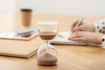 Wall Mural - Hourglass with flowing sand on desk. Woman taking notes while using calculator indoors, selective focus