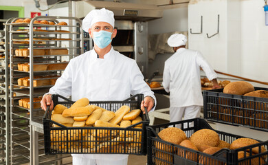 Wall Mural - Baker in face mask for viral protection holding crate with bread, industrial kitchen of bakery on background