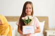 © Pixel-Shot - Little girl with greeting card for Mother's Day and tulips in bedroom