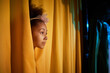 Side view portrait of African American girl peeking head on stage from yellow curtains with spotlight, copy space