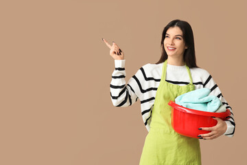 Sticker - Beautiful young woman holding basket with clean laundry and pointing at something on brown background