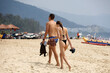Young couple in swimwear walking by white sand beach on background of mountains. Man and slim girl in bikini together, romantic leisure on tropical coast