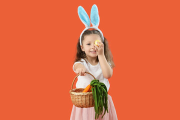 Sticker - Cute little girl in bunny ears holding wicker basket with toys carrots and Easter egg on orange background