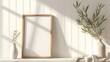 Blank vertical wooden poster frame standing mockup on a white wall near a white vase of olive leaves and beige fabric over a white table