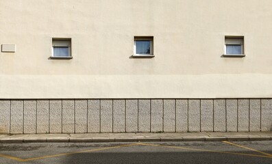 Row of small windows on pale yellow plaster facade with concrete blocks below. Sidewalk and urban street in front. Background for copy space.