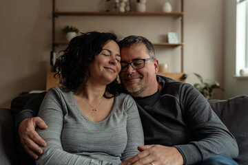 A middle-aged couple, both with light skin, cuddles on a couch in a cozy living room. The woman, with curly hair, smiles with her eyes closed, leaning on the man wearing glasses and smiling