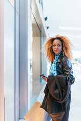 Canvas Print - Young black girl looks at her mobile phone in front of the window of a store inside a shopping center